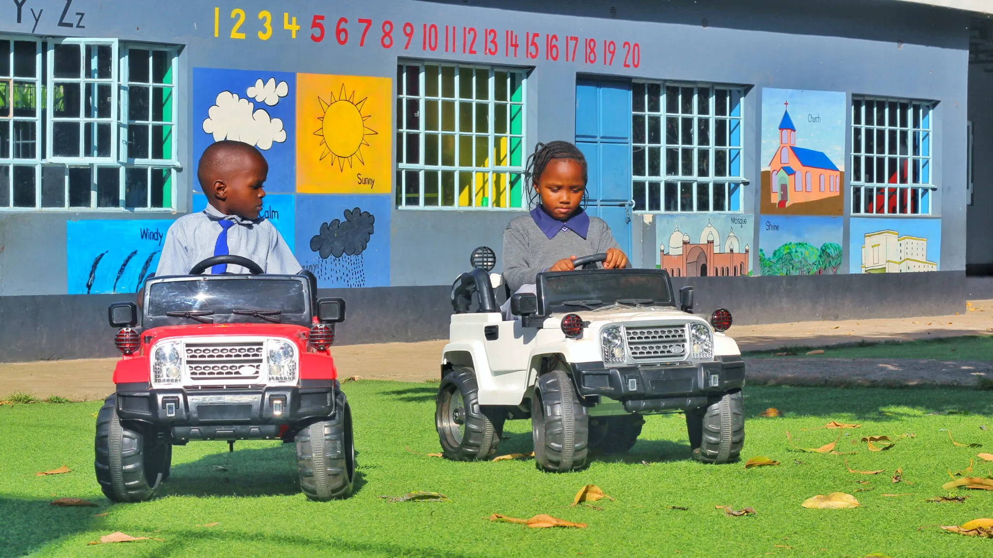 Two small children driving toy jeeps in front of a painted classroom wall