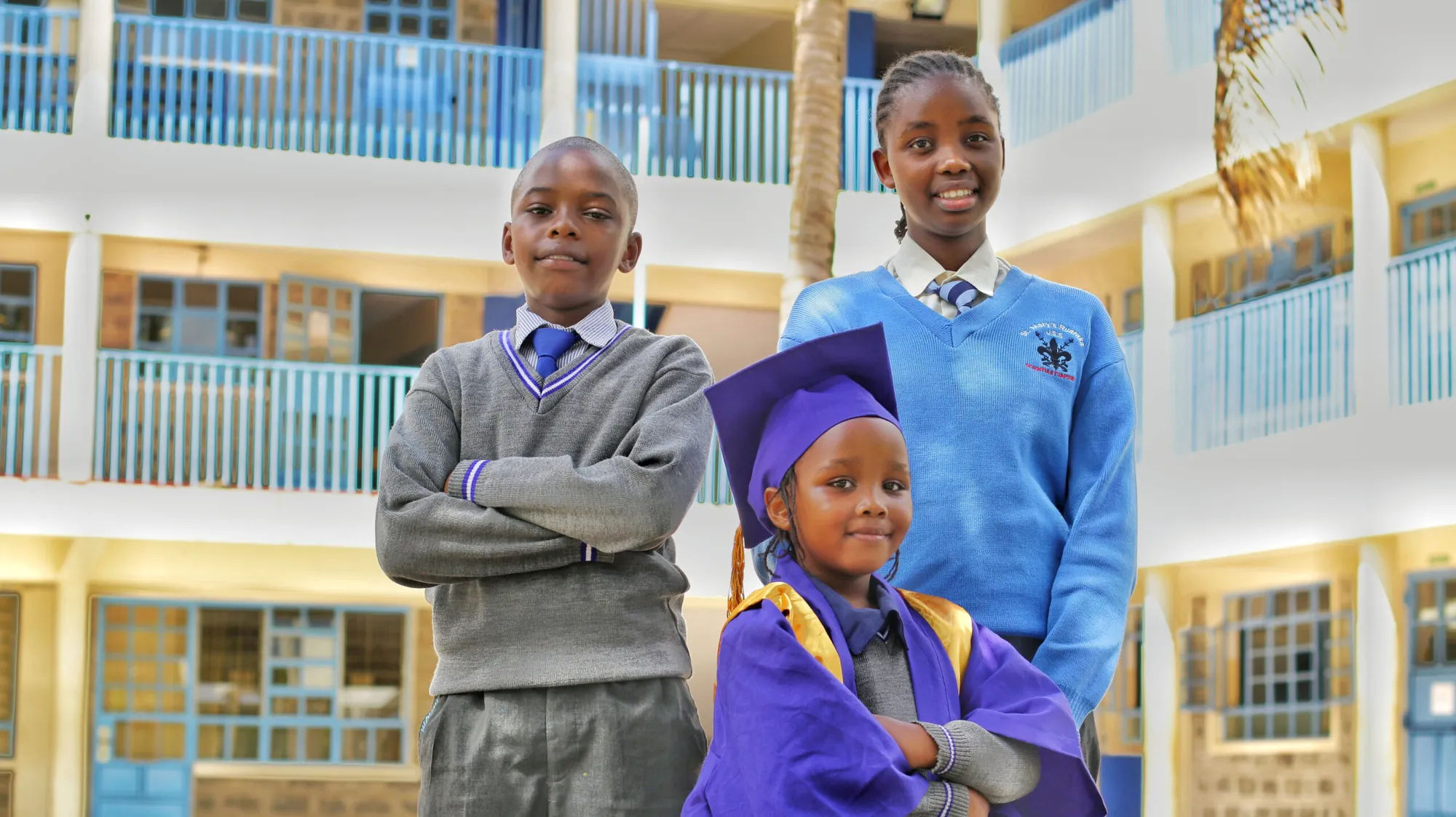 Three pupils in school uniform and graduation attire posing in front of the school building
