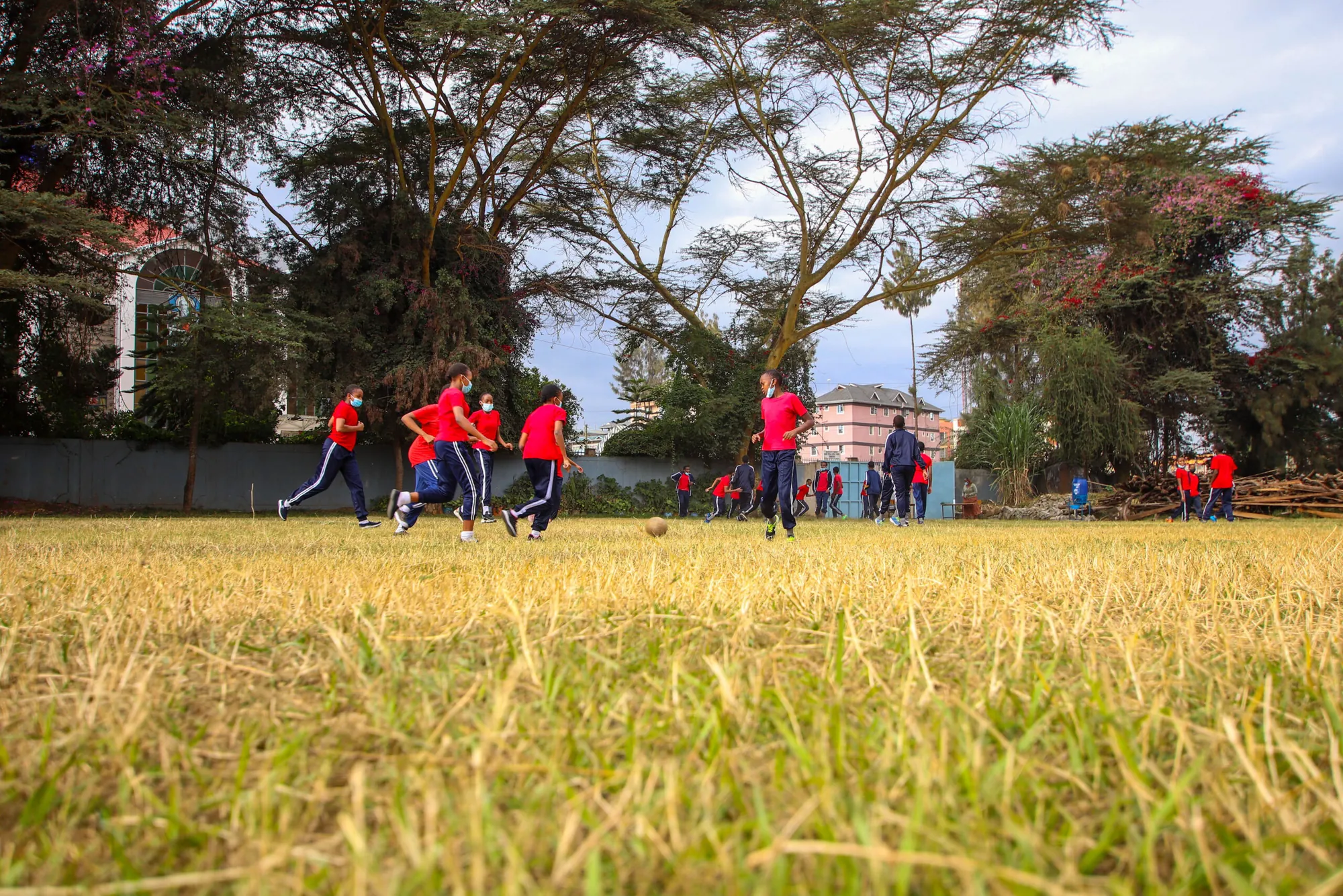 Pupils in red running across a dry grass field under tall trees