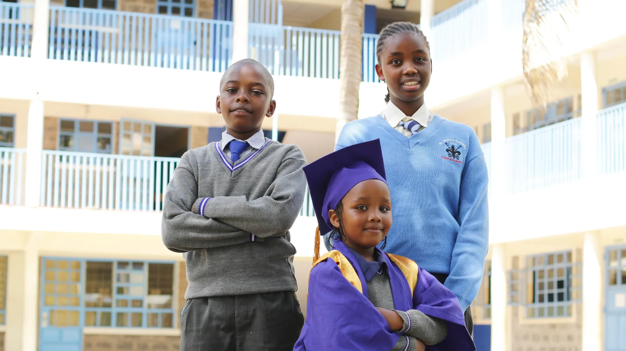 Three pupils in school uniform and graduation attire posing in front of the school building